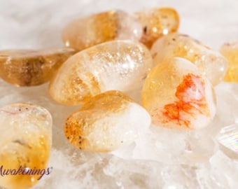 May include: A collection of tumbled citrine stones, featuring a variety of shapes and sizes. The stones are a light yellow color with some orange and red hues. The stones are arranged on a white surface.
