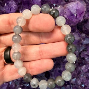 May include: A beaded bracelet featuring a mix of gray and white round gemstones. The bracelet is held in a hand, showcasing the varying shades of the beads. The background is a cluster of purple crystals.