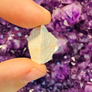 Puede incluir: Un pequeño cristal blanco, rugoso y texturizado, con una punta puntiaguda, sostenido en una mano sobre un fondo de cristales de amatista púrpura.