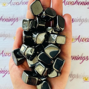 May include: A pile of black hematite tumbled stones in a hand. The stones are smooth and shiny.