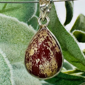 May include: A teardrop-shaped pendant with a silver-colored setting. The pendant features a polished stone with a maroon and beige mottled pattern. The bail is a simple, decorative design. The background is a blurred green.