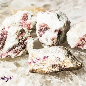 May include: Five rough, white and pink, raw, natural, tumbled, pink tourmaline crystals. The crystals are clustered together on a white, textured surface.