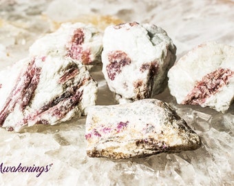 Puede incluir: Cinco cristales de turmalina rosa en bruto, de color blanco y rosa, naturales, pulidos y redondeados. Los cristales están agrupados sobre una superficie blanca texturizada.