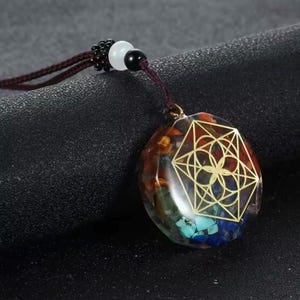 Könnte beinhalten: Eine Anhängerkette mit einem goldenen geometrischen Design über einem farbenfrohen Stein. Der Stein ist eine Mischung aus roten, blauen, grünen und gelben Fragmenten. Das Halsband ist dunkelbraun mit schwarzen und weißen Perlen.