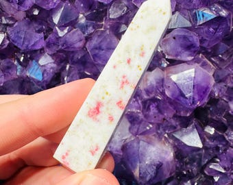Puede incluir: Una punta de cristal blanca y rosa con una textura rugosa. El cristal está sentado en un lecho de cristales de amatista púrpura.
