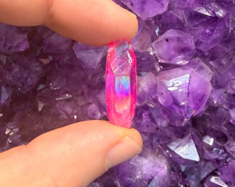May include: A small, pink crystal with iridescent highlights is held between two fingers against a background of purple amethyst crystals. The crystal is a vibrant pink color with a pointed tip and a textured surface. The background is a cluster of deep purple amethyst crystals.
