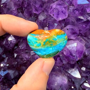 Puede incluir: Una piedra pulida en forma de corazón con remolinos vibrantes de azul turquesa, verde intenso y marrón anaranjado cálido. La superficie lisa y redondeada muestra un patrón jaspeado con transiciones de color claras en todo el objeto.