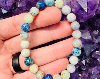 Puede incluir: Un brazalete hecho de cuentas de piedras preciosas azules, verdes y blancas. El brazalete está en una mano, y el fondo es un montón de cristales de amatista púrpura.