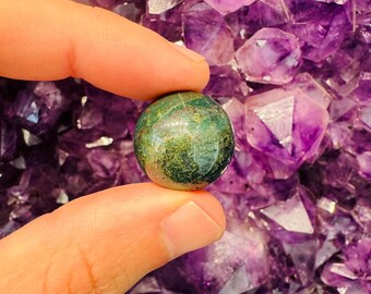 May include: A small, round, green and brown gemstone sphere is held between two fingers. The background is a cluster of purple amethyst crystals, creating a contrast in color and texture. The sphere appears to be polished.