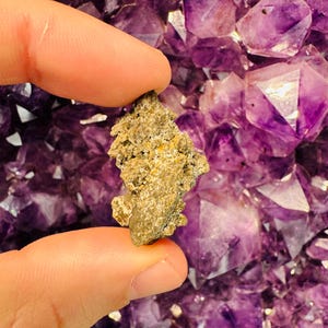 Puede incluir: Una pequeña roca rugosa de color gris-marrón con una punta puntiaguda, sostenida en una mano sobre un fondo de cristales de amatista púrpura.