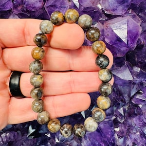 May include: A beaded bracelet featuring round, polished stones in shades of brown, gray, and gold. The bracelet is held against a background of purple crystals, showcasing the natural variations in the stone's color and texture. The bracelet is a piece of jewelry.