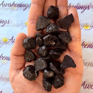 Pode incluir: Um punhado de pedras de obsidiana pretas e &aacute;speras. As pedras t&ecirc;m formas irregulares e uma superf&iacute;cie brilhante e reflexiva. O fundo apresenta a palavra "Awakenings".