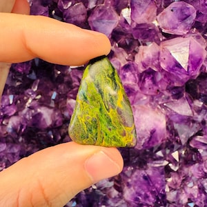 Puede incluir: Una piedra verde y amarilla con patrones en espiral, sobre un fondo de cristales de amatista púrpura.