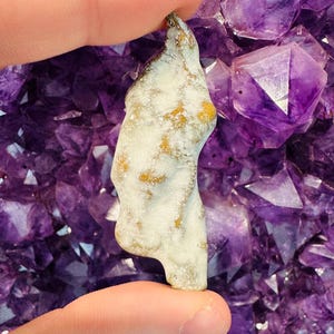 May include: A small, white and yellow, irregularly shaped rock with a rough texture. The rock is being held by a hand against a background of purple amethyst crystals.