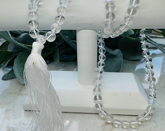 Op de afbeelding: Een ketting van heldere kwarts kristallen kralen met een witte kwast. De ketting wordt getoond op een witte displaystandaard. De ronde, transparante kralen zijn geregen en de kwast is gemaakt van witte draden.