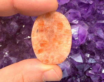 May include: An oval-shaped, orange sunstone gemstone with a speckled pattern is held between two fingers. The background is a cluster of deep purple amethyst crystals.