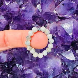 May include: A beaded ring composed of small, round, off-white and gray stones. A single silver bead is centered on the ring. The ring is displayed against a background of purple amethyst crystals.