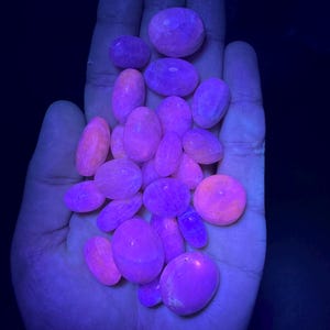 May include: A collection of smooth, oval-shaped stones in shades of pink and purple, illuminated under a UV light, showcasing their vibrant fluorescence.
