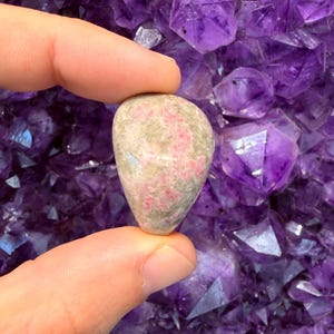May include: A polished, teardrop-shaped stone featuring a mottled surface with patches of vibrant pink and muted grey. The stone has a smooth, rounded texture and exhibits natural color variations throughout its compact, solid form.