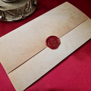 May include: A vintage-style envelope with a red wax seal. The envelope is made of aged, tan-colored paper with a darker, faded border. The seal is circular, with a detailed design. The envelope is on a red surface.
