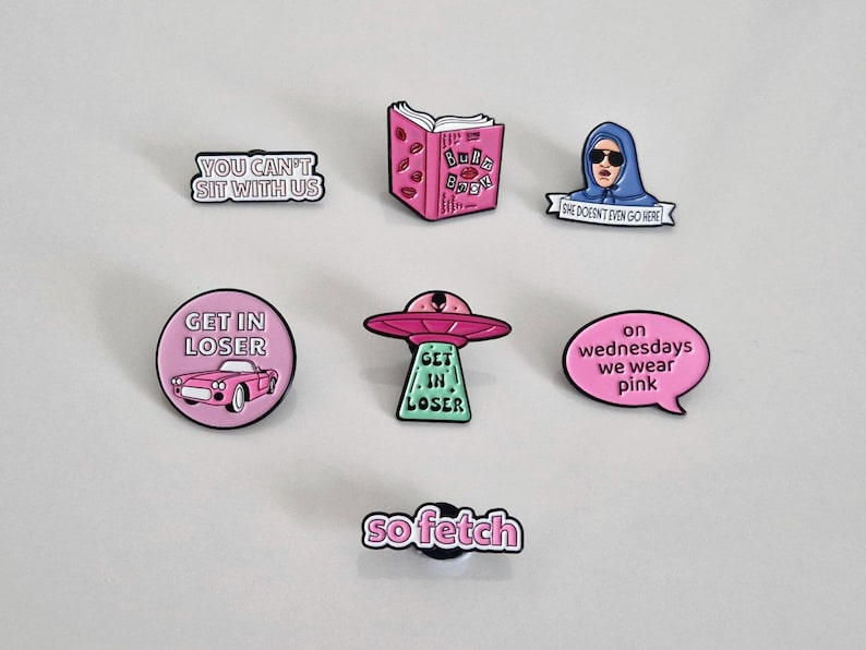 Puede incluir: Diez pines de esmalte rosa con diferentes lemas e im&aacute;genes. Los pines incluyen "You Can't Sit With Us", "Get In Loser", "Get In Loser", "On Wednesdays We Wear Pink", "She Doesn't Even Go Here", "So Fetch", y un libro rosa con las palabras "Burn Book" en la portada.