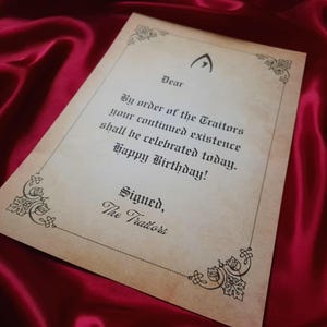 May include: A parchment-style birthday card with ornate corner designs and the text "By order of the Traitors your continued existence shall be celebrated today. Happy Birthday!" on a red satin background.