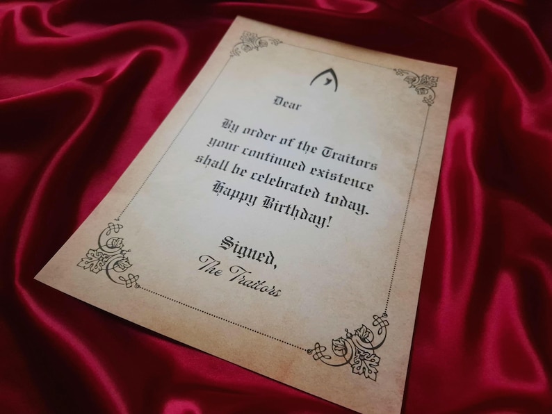 May include: A parchment-style birthday card with ornate corner designs and the text "By order of the Traitors your continued existence shall be celebrated today. Happy Birthday!" on a red satin background.