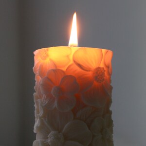 K&ouml;nnte beinhalten: Eine wei&szlig;e Kerze mit floralem Design und einem brennenden Docht. Die Kerze ist blumenf&ouml;rmig und hat einen warmen, orangenen Schein.