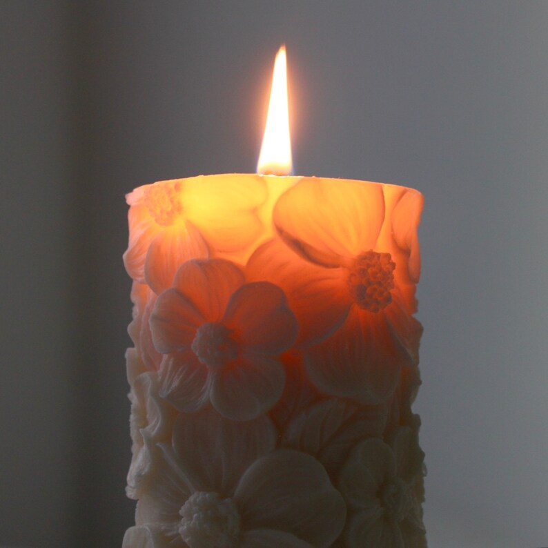 K&ouml;nnte beinhalten: Eine wei&szlig;e Kerze mit floralem Design und einem brennenden Docht. Die Kerze ist blumenf&ouml;rmig und hat einen warmen, orangenen Schein.