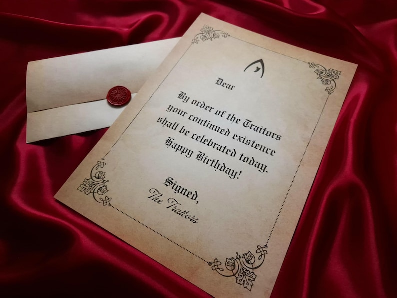 May include: A parchment-style birthday card with an envelope, sealed with a red wax stamp, rests on a deep red satin surface. The card features ornate borders and gothic-style text that reads "By order of the Traitors...Happy Birthday!"