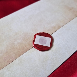 May include: An envelope with a red wax seal. The envelope is off-white and tan, with a diagonal seam. The seal is red with a small white square in the center.