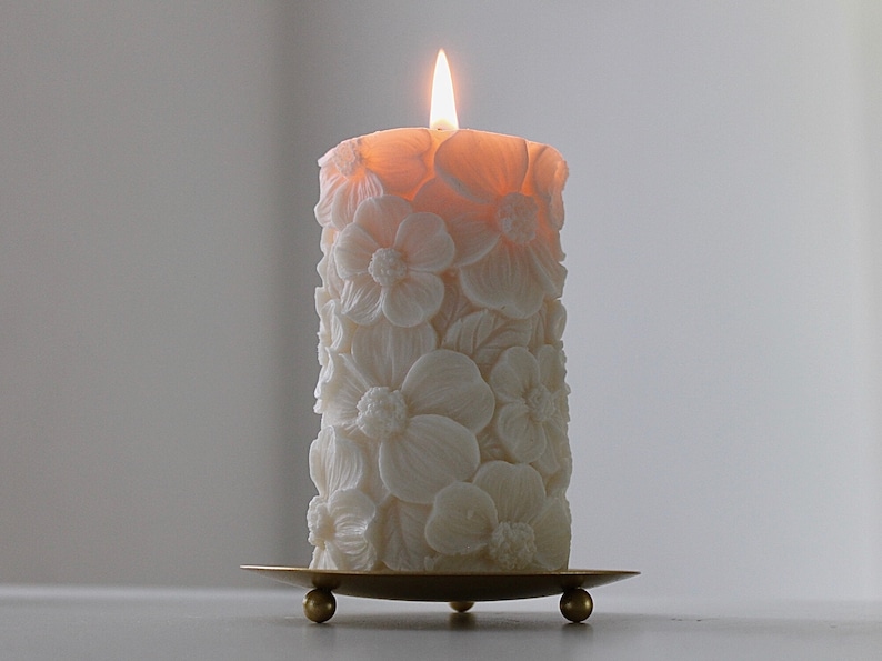 K&ouml;nnte beinhalten: Eine wei&szlig;e Kerze mit floralem Design und einer brennenden Flamme. Die Kerze steht auf einem goldenen Metallst&auml;nder mit drei Beinen.