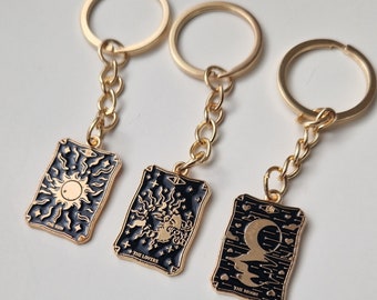 Könnte beinhalten: Drei goldene Schlüsselanhänger mit schwarzen Emaille-Anhängern mit Tarotkarten-Motiven. Die Anhänger zeigen die Karten "Die Sonne", "Die Liebenden" und "Der Mond".