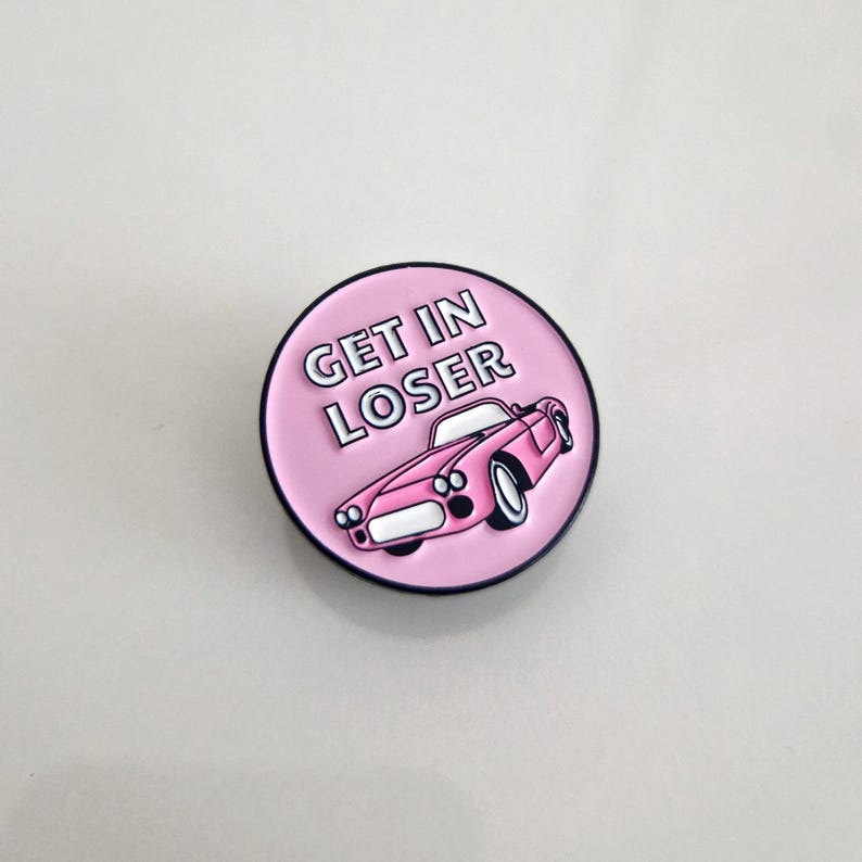 Puede incluir: Pin de esmalte rosa con contorno blanco. El pin presenta un coche rosa con el texto "Get in Loser" en letras blancas.