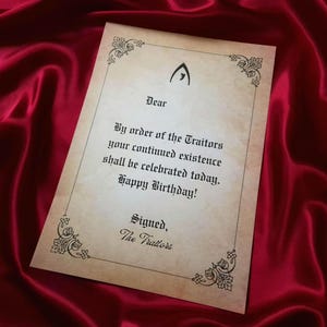 May include: A parchment-style birthday card with ornate corner designs and the text "By order of the Traitors your continued existence shall be celebrated today. Happy Birthday!" on a red satin background.