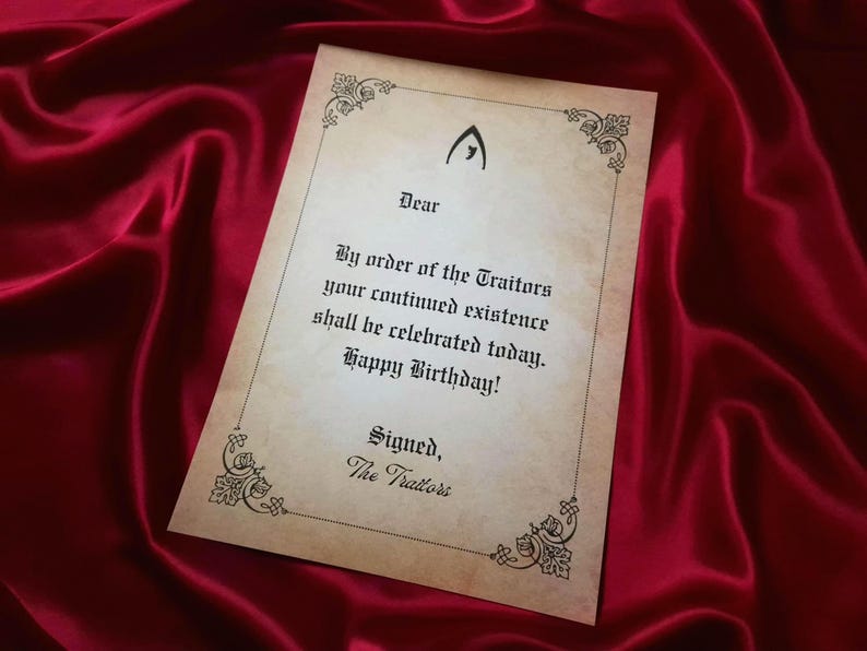 May include: A parchment-style birthday card with ornate corner designs and the text "By order of the Traitors your continued existence shall be celebrated today. Happy Birthday!" on a red satin background.