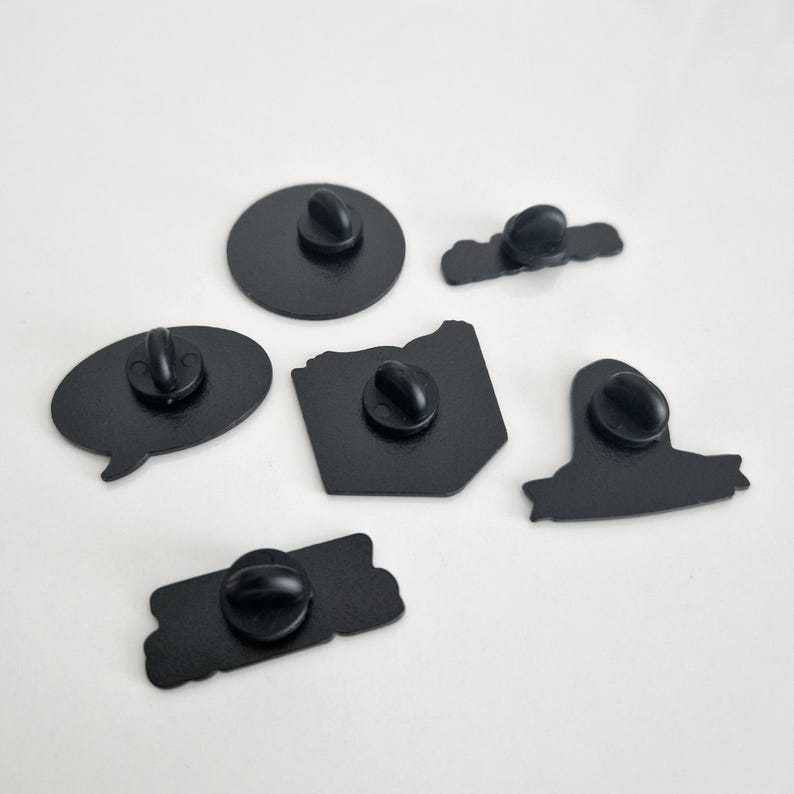 Puede incluir: Seis pines de esmalte negro con diferentes dise&ntilde;os. Las pines tienen un di&aacute;metro de aproximadamente 5 cm y tienen un respaldo de metal plateado.