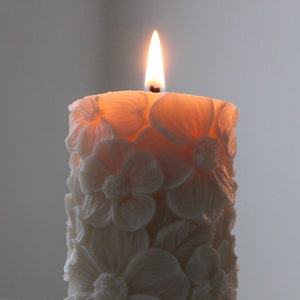 K&ouml;nnte beinhalten: Eine wei&szlig;e Kerze mit Blumenmuster ist angez&uuml;ndet und brennt. Die Kerze ist aus Wachs und hat ein zartes, filigranes Blumendesign.