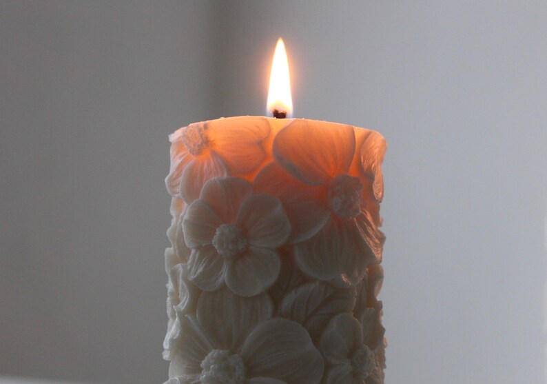 K&ouml;nnte beinhalten: Eine wei&szlig;e Kerze mit Blumenmuster ist angez&uuml;ndet und brennt. Die Kerze ist aus Wachs und hat ein zartes, filigranes Blumendesign.
