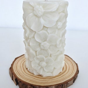 K&ouml;nnte beinhalten: Wei&szlig;e Kerze in S&auml;ulenform mit floralem Design. Die Kerze steht auf einer Holzscheibe.