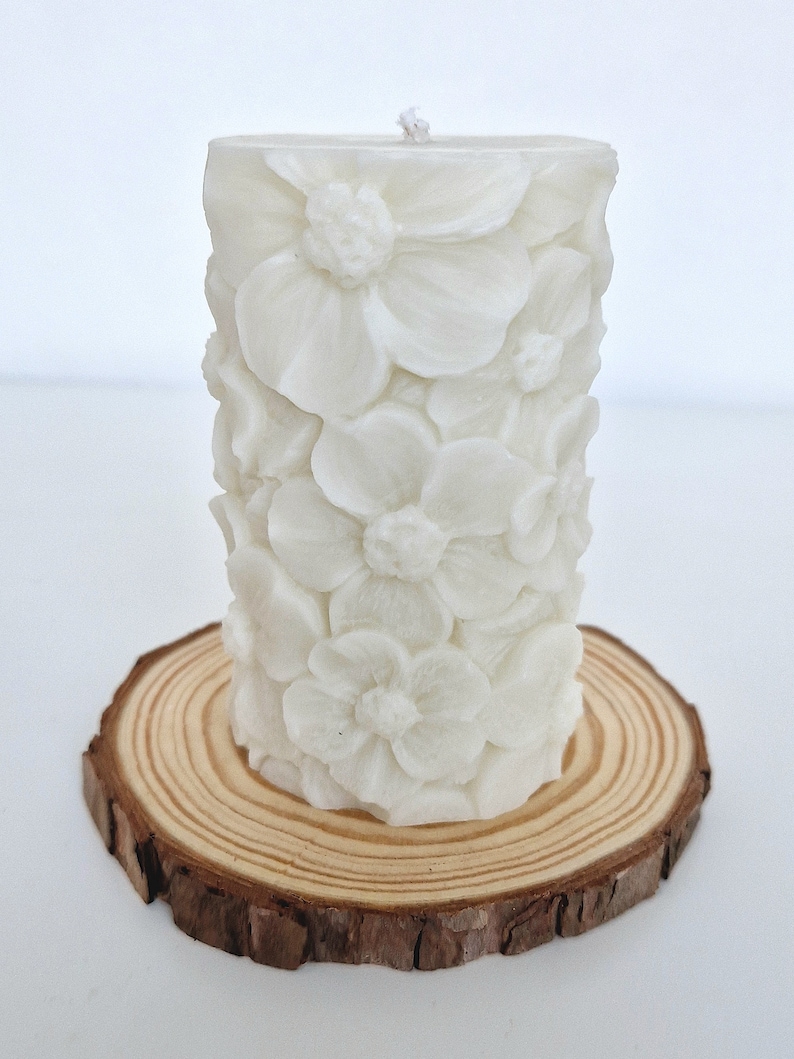 K&ouml;nnte beinhalten: Wei&szlig;e Kerze in S&auml;ulenform mit floralem Design. Die Kerze steht auf einer Holzscheibe.