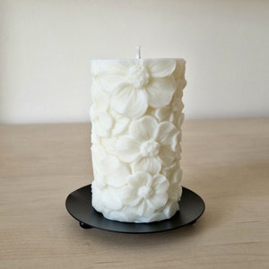 K&ouml;nnte beinhalten: Wei&szlig;e Kerze in S&auml;ulenform mit Blumenmuster. Die Kerze steht auf einem schwarzen Kerzenhalter.