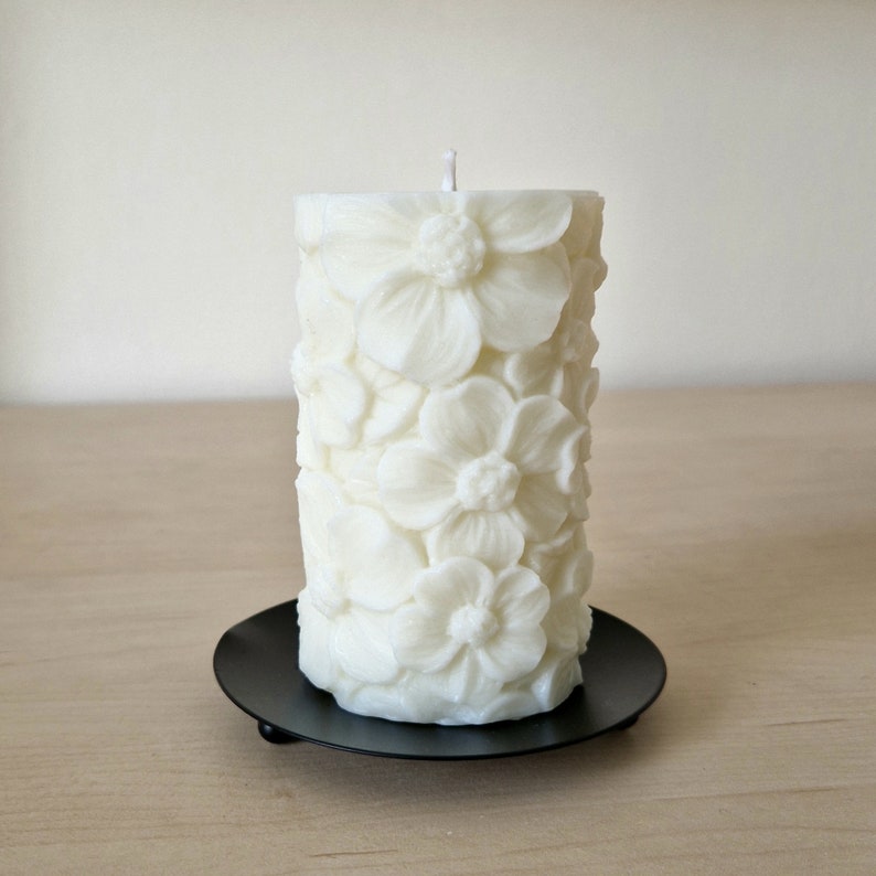K&ouml;nnte beinhalten: Wei&szlig;e Kerze in S&auml;ulenform mit Blumenmuster. Die Kerze steht auf einem schwarzen Kerzenhalter.