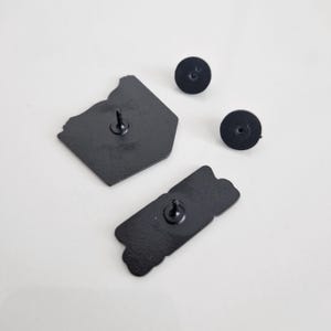 Puede incluir: Vista posterior de dos pines de esmalte negro. Un pin tiene forma de contenedor para llevar y el otro es una pancarta rectangular con la palabra "GOOD" escrita en ella. Ambos pines tienen un respaldo de metal negro.