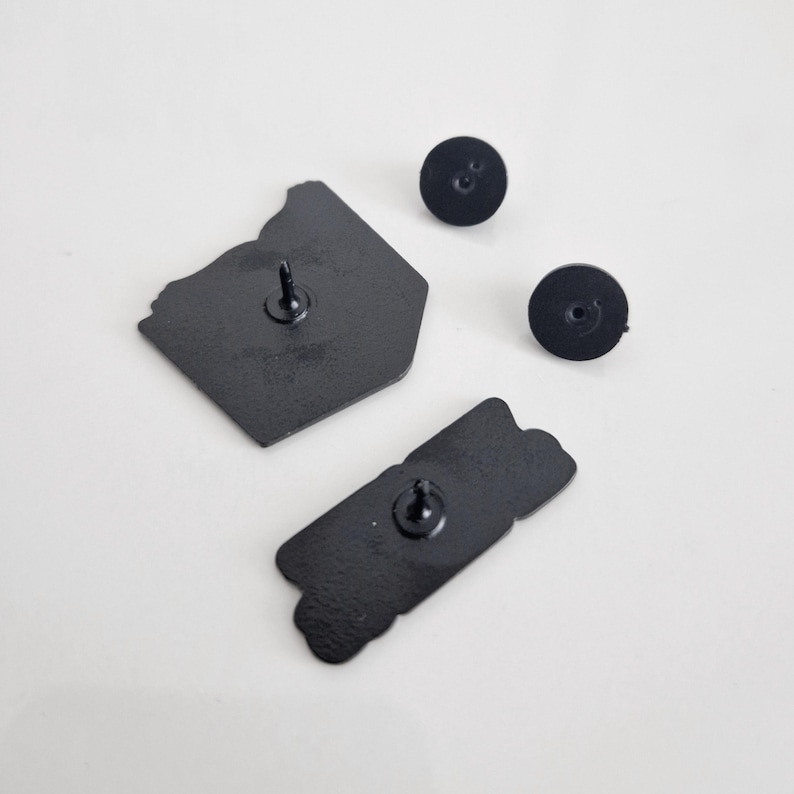 Puede incluir: Vista posterior de dos pines de esmalte negro. Un pin tiene forma de contenedor para llevar y el otro es una pancarta rectangular con la palabra "GOOD" escrita en ella. Ambos pines tienen un respaldo de metal negro.