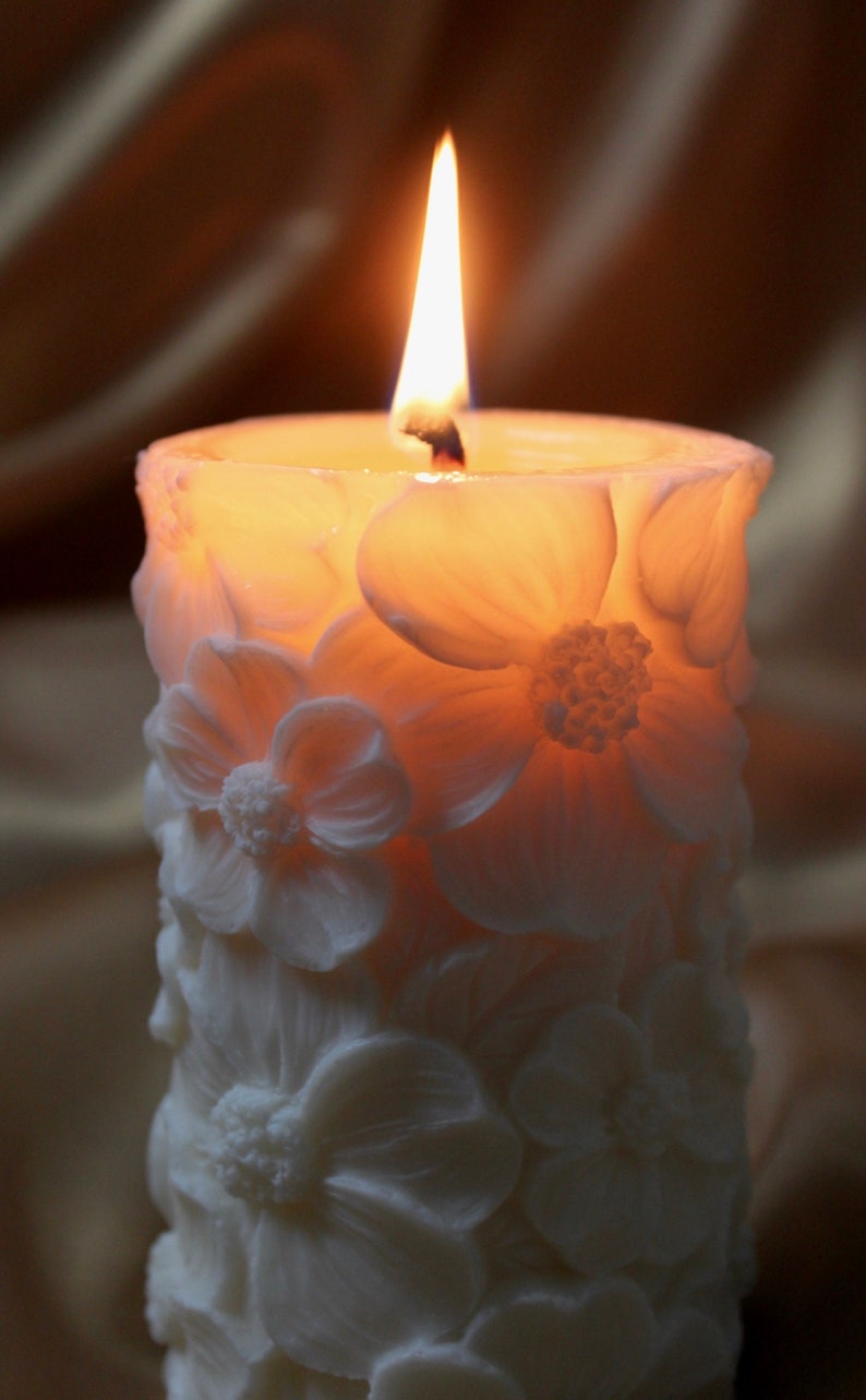K&ouml;nnte beinhalten: Eine wei&szlig;e Kerze mit floralem Design und einem brennenden Docht. Die Kerze ist aus Wachs und hat ein zartes, weibliches Aussehen.