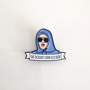 Puede incluir: Un pin de esmalte azul con un personaje de dibujos animados que lleva una sudadera con capucha azul y gafas de sol. El personaje sostiene una pancarta que dice "She Doesn't Even Go Here."