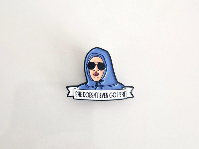 Puede incluir: Un pin de esmalte azul con un personaje de dibujos animados que lleva una sudadera con capucha azul y gafas de sol. El personaje sostiene una pancarta que dice "She Doesn't Even Go Here."