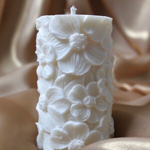 K&ouml;nnte beinhalten: Eine wei&szlig;e Kerze in S&auml;ulenform mit einem floralen Design. Die Kerze ist mit erhabenen wei&szlig;en Blumen verziert.