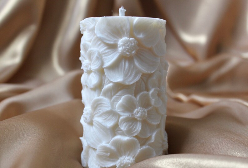 K&ouml;nnte beinhalten: Eine wei&szlig;e Kerze in S&auml;ulenform mit einem floralen Design. Die Kerze ist mit erhabenen wei&szlig;en Blumen verziert.